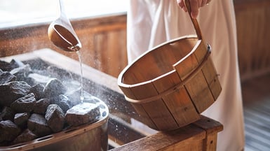 Eine Person mit umgebundenen Handtuch kippt Wasser auf heiße Steine in einer Sauna